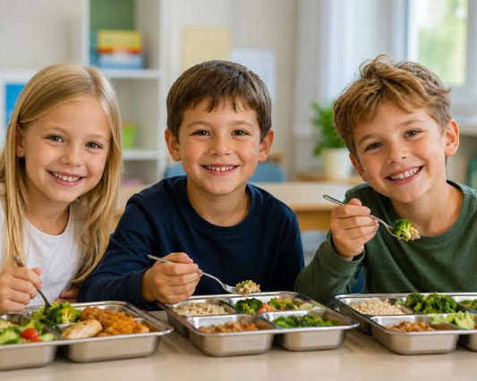 Three smiling children sitting at a table with metal meal trays in front of them, holding forks in a bright indoor setting.