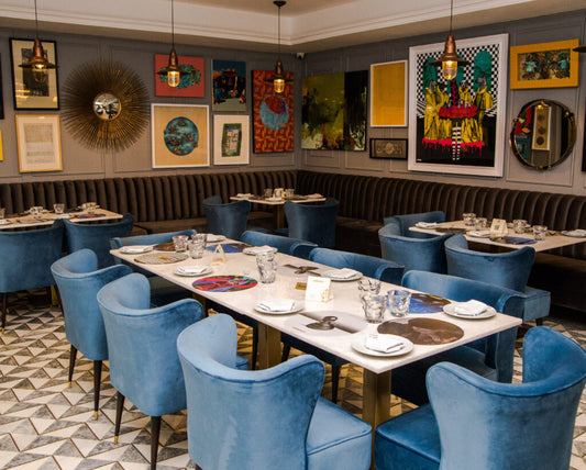 A styled restaurant dining room with multiple white rectangular tables set with plates and glasses, surrounded by blue velvet chairs. The room features dark brown tufted banquette seating, gray walls decorated with numerous colorful framed artworks, and hanging pendant lights.