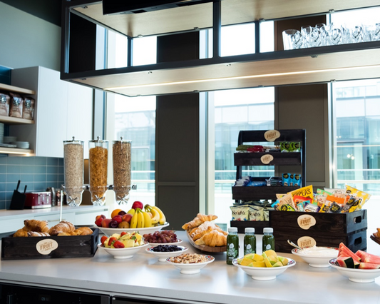 A breakfast buffet spread on a white counter in a modern kitchen or cafe setting, with breads, croissants, fruit, yogurt, cereals, and bottled drinks in front of large windows.